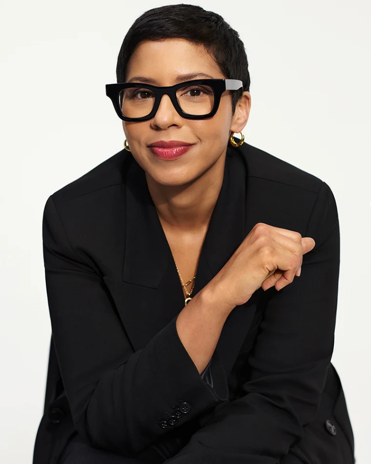 Person with short hair and glasses wearing a black blazer, gold jewelry, and smiling slightly against a plain background.