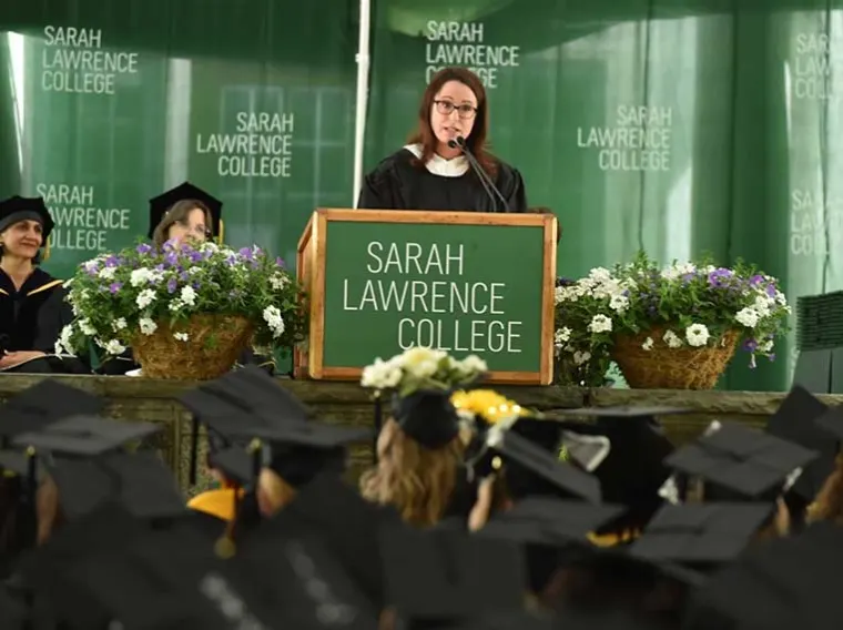 Person speaking at a podium with "Sarah Lawrence College" text. Audience in graduation caps. Flowers and college banner in the background.