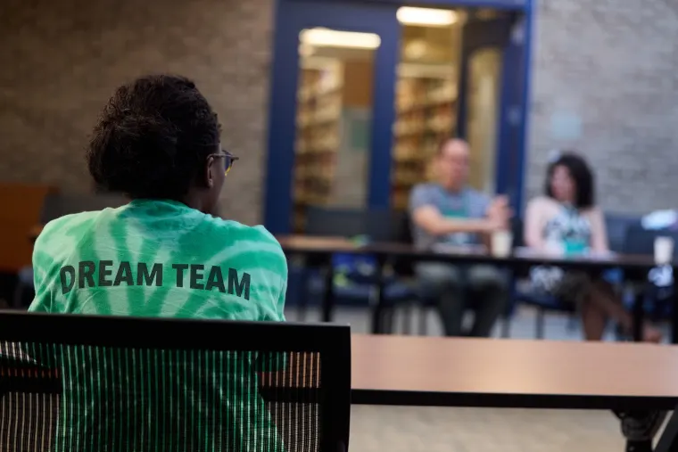 Person in green "Dream Team" shirt sits facing a table, with two others blurred in the background.