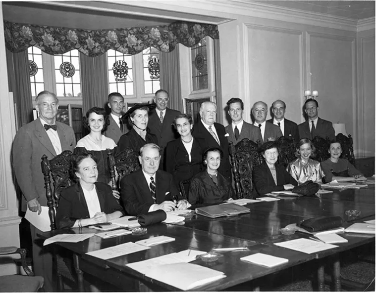 A large group of people in formal attire is seated and standing around a table in an ornate room with papers spread in front.