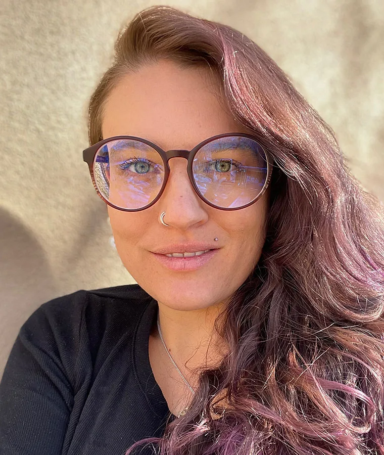 Person with long, wavy hair, wearing glasses, a nose ring, and a lip stud, smiling slightly.