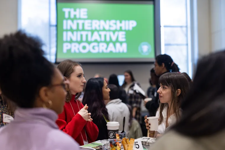 People converse at tables in a room. A screen in the background displays the text "The Internship Initiative Program."