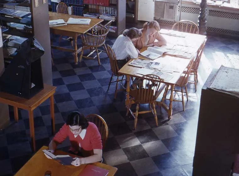 Library scene with two people reading at adjacent tables, surrounded by bookshelves. Sunlight streams in through a window.