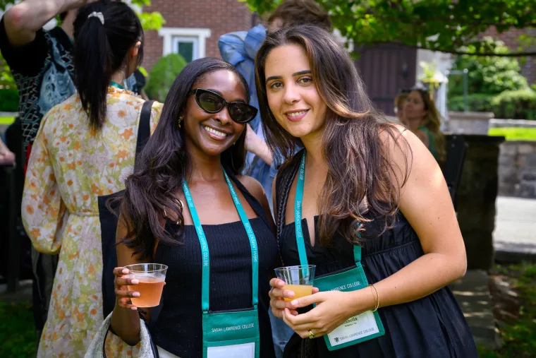 Two people outdoors in summer attire with lanyards, holding drinks, smiling at the camera. Others are in the background.
