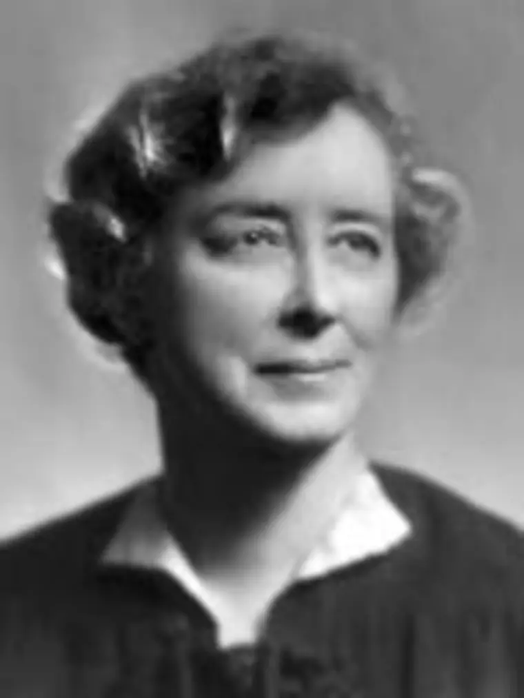 Person with short curly hair wearing a dark top and white collar looking slightly to the side against a neutral background.