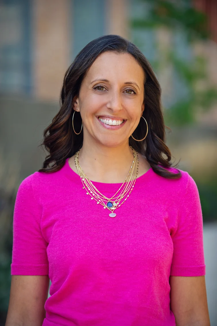 Person smiling, wearing a bright pink top and layered gold necklaces, standing outdoors with a blurred background.