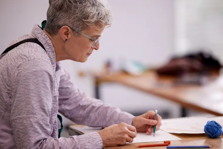 Person with short gray hair, wearing glasses and a light jacket, writing on paper at a desk with a blue yarn ball and orange pen nearby.