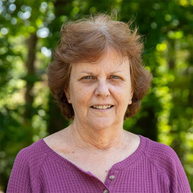 Person with short brown hair wearing a purple top, smiling outdoors with green trees blurred in the background.