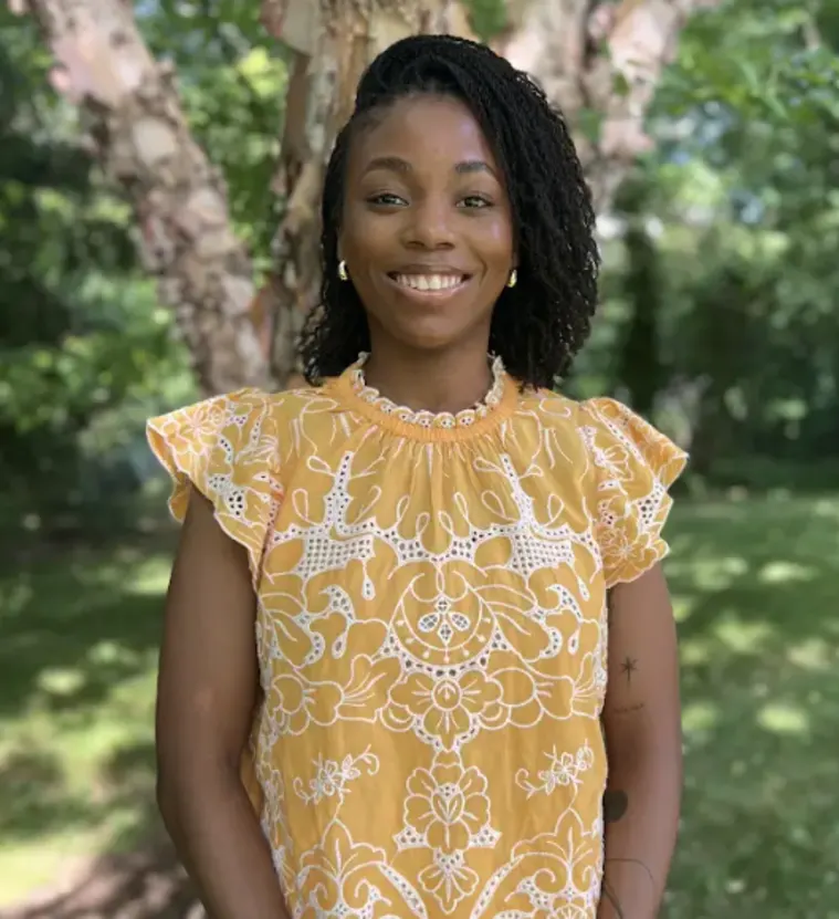 Person smiling outdoors wearing a yellow patterned dress, standing in front of a tree with green foliage in the background.