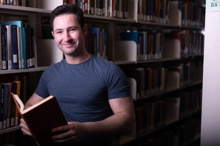 Person smiling while holding an open book, standing in a library aisle with shelves full of books.