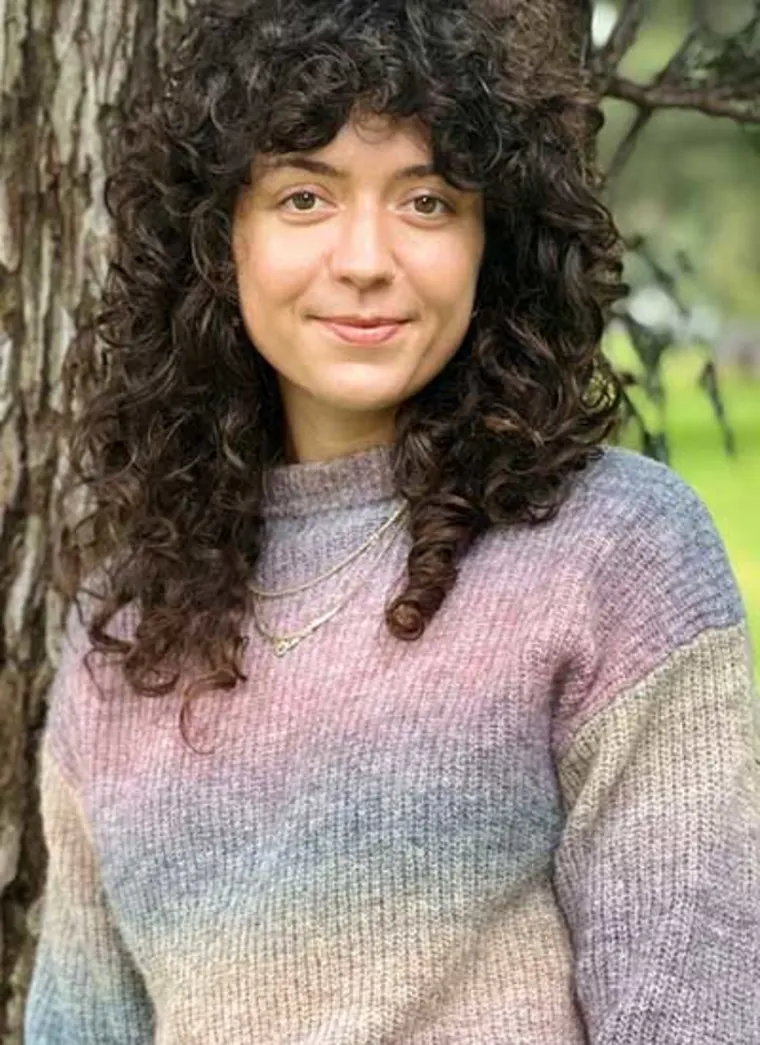 Person with curly hair smiling, wearing a colorful sweater and standing by a tree in a park.