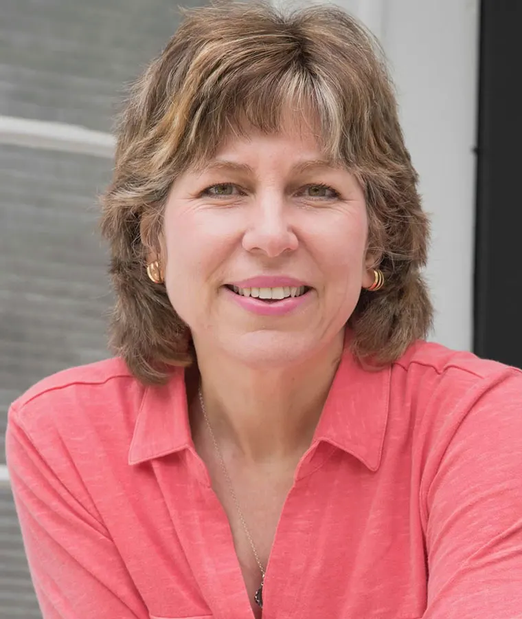 Person smiling, wearing a coral shirt and gold earrings, with short brown hair, standing in front of a window.