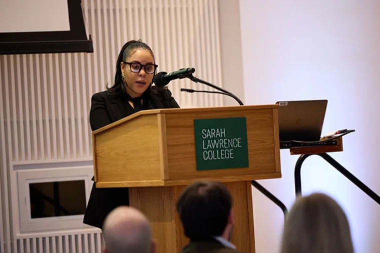 Person speaking at a wooden podium with a "Sarah Lawrence College" sign, addressing an audience, with a laptop and microphone nearby.