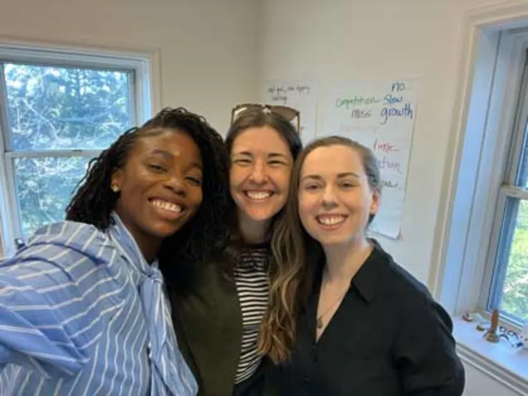 Three people smiling indoors, standing close together. There's sunlight coming through a window and notes on a whiteboard in the background.