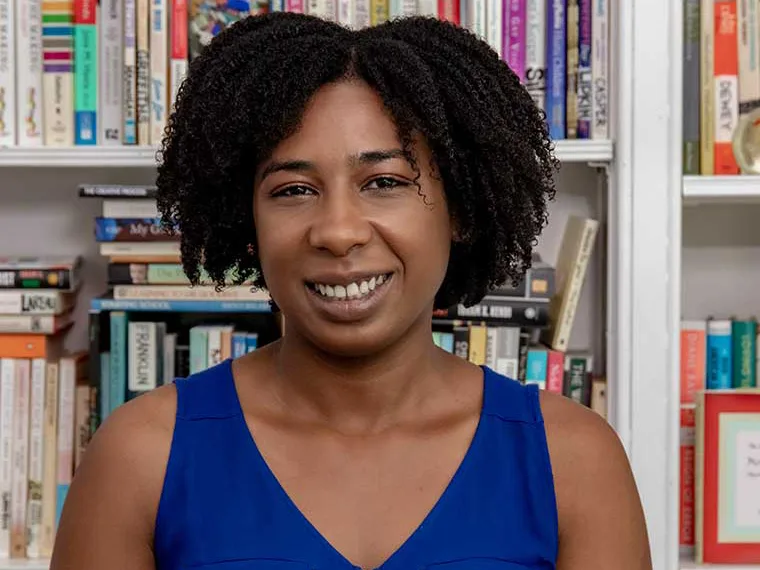 Person with curly hair smiles in front of a bookshelf filled with various books, wearing a blue top.
