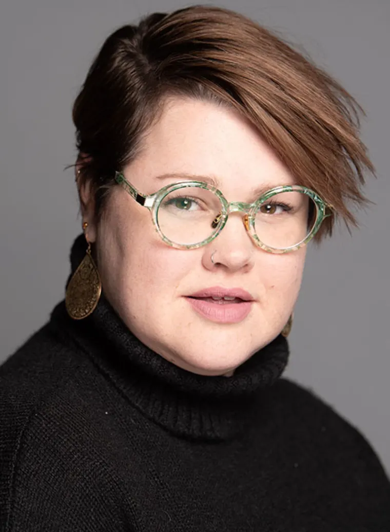 Person with short brown hair, round glasses, nose ring, and large earrings, wearing a black turtleneck, against a gray background.