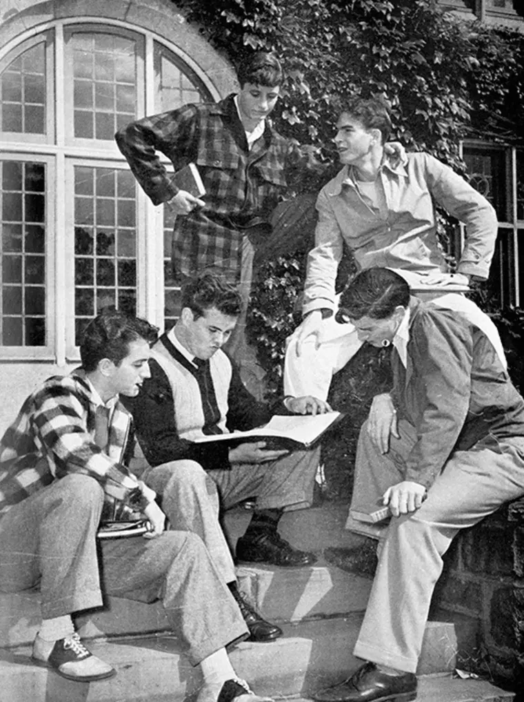 Five people in casual clothing sit and stand on stairs outside a building with ivy, looking at a book.