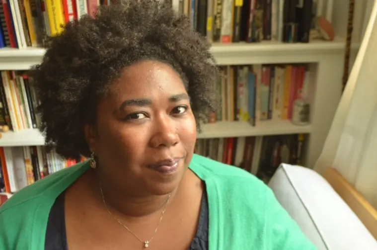 Person with curly hair in a green sweater, sitting in a room with a bookshelf in the background.