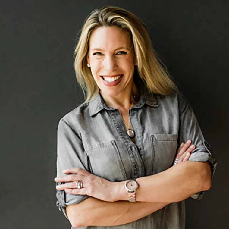 Person with long hair smiles, arms crossed, wearing a gray shirt and watch, standing against a dark background.