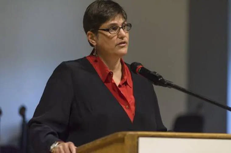 Person with short hair and glasses speaks at a podium, wearing a black jacket and red shirt, with a microphone visible.