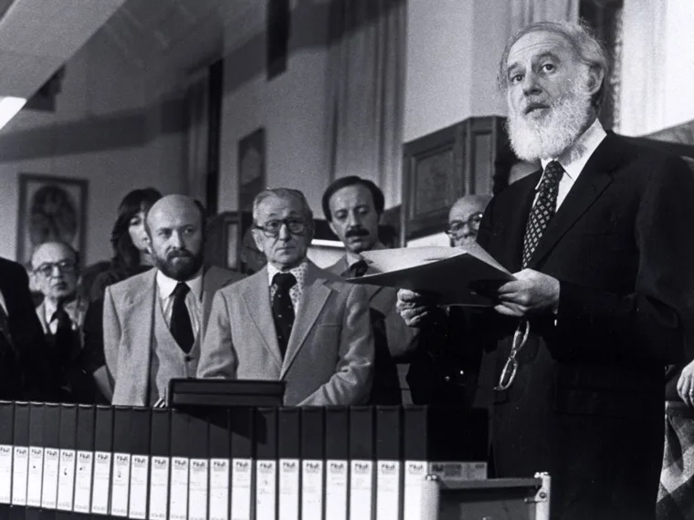 A group of people in formal attire stands behind a speaker holding papers, with binders lined up in the foreground.