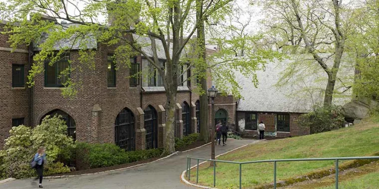 Brick building with large windows, surrounded by trees and grass. A few people walk along the path in front of the building.