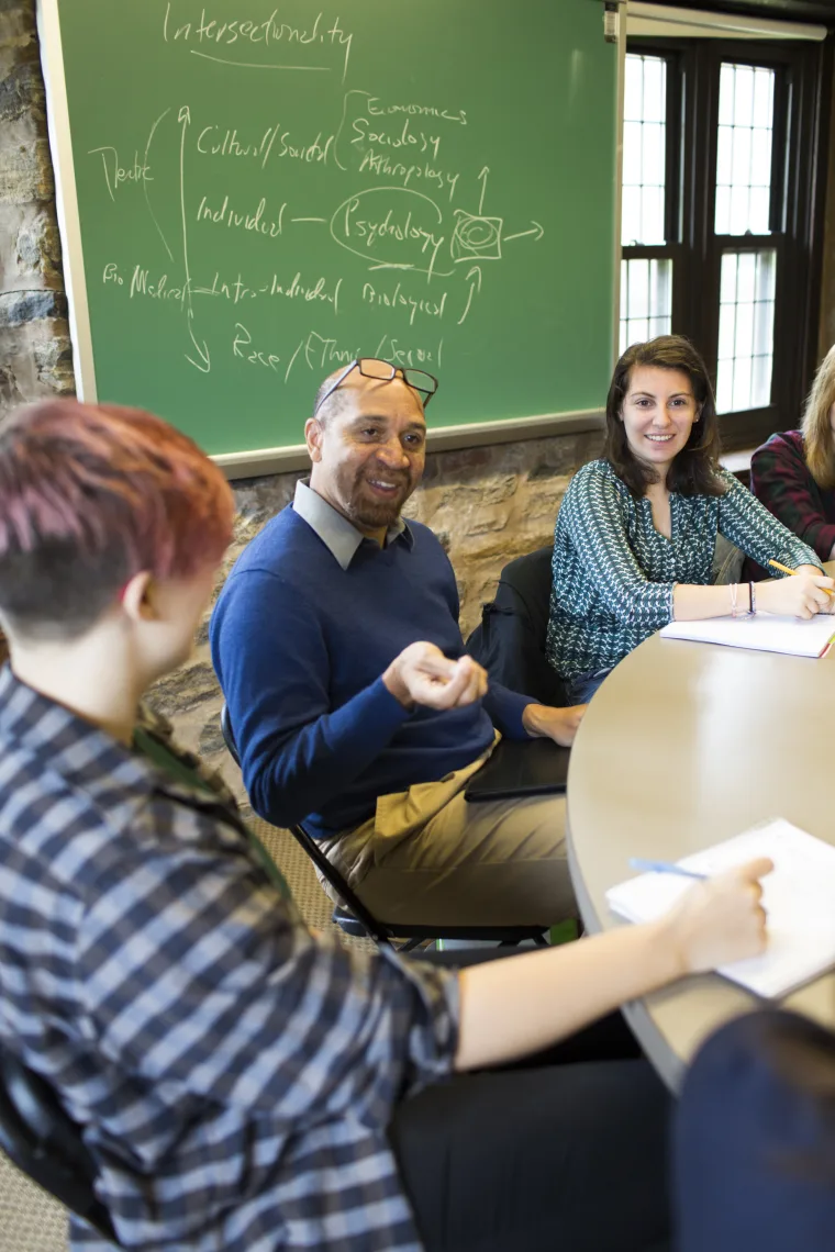 Three people having a discussion around a table, with a chalkboard in the background displaying handwritten notes and diagrams.