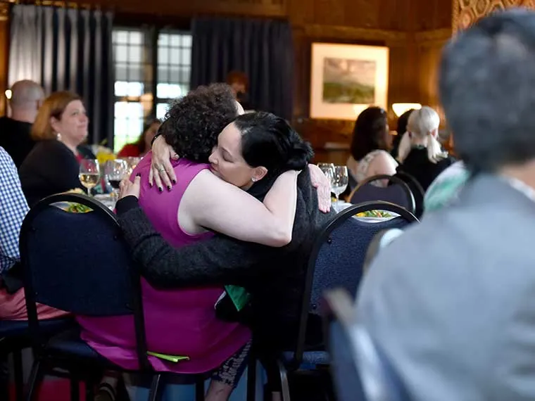 Two people hugging at an event surrounded by seated attendees and tables with glasses.