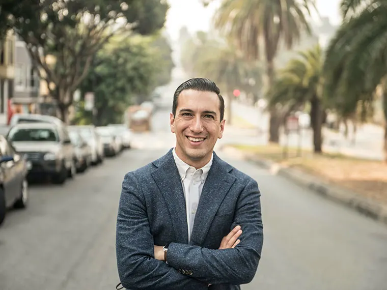 Person smiling with arms crossed, standing on a palm-lined street.