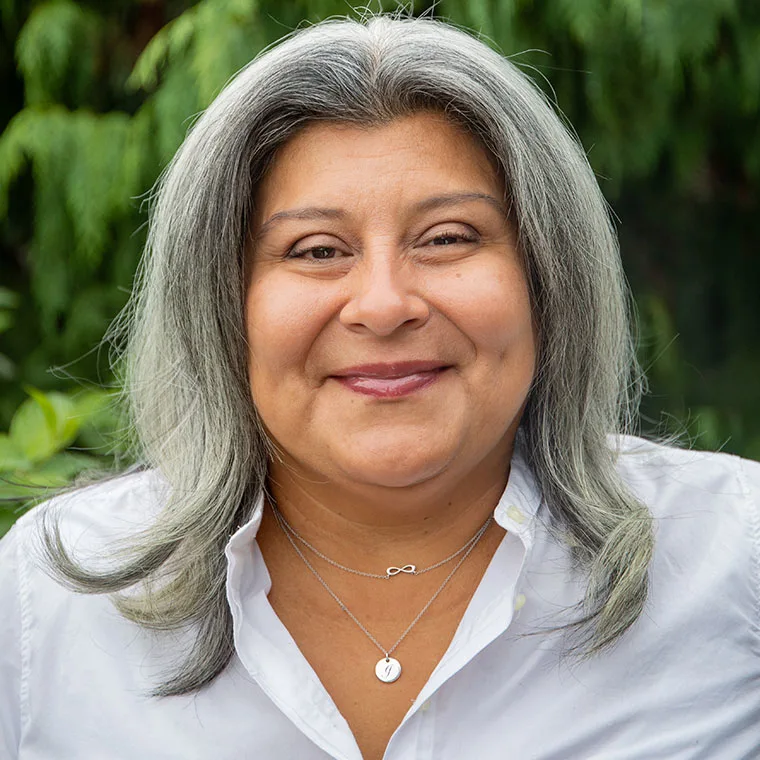 Person with gray hair smiling, wearing a white shirt and two necklaces. Greenery in the blurred background.