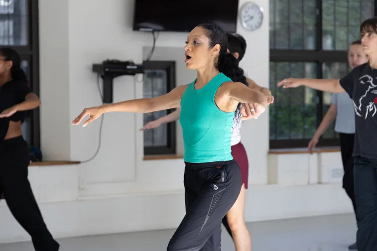 Person in a turquoise top and black pants leads a dance class, arms extended. Others follow, mirroring the pose in a well-lit studio.