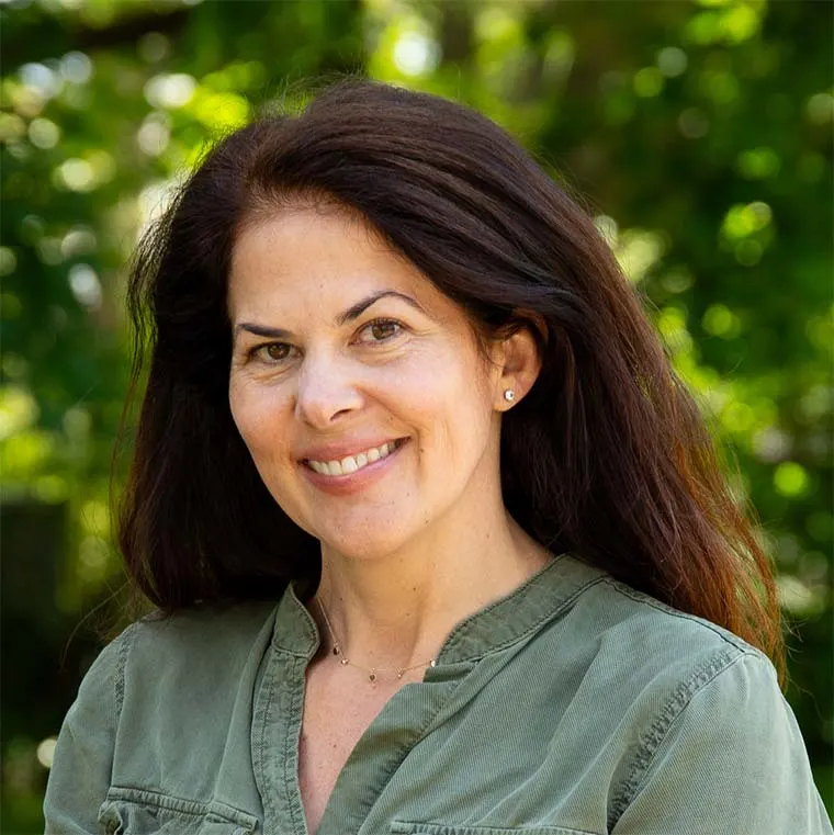 A person with long dark hair smiles, wearing a green shirt, standing outdoors with blurred green foliage in the background.