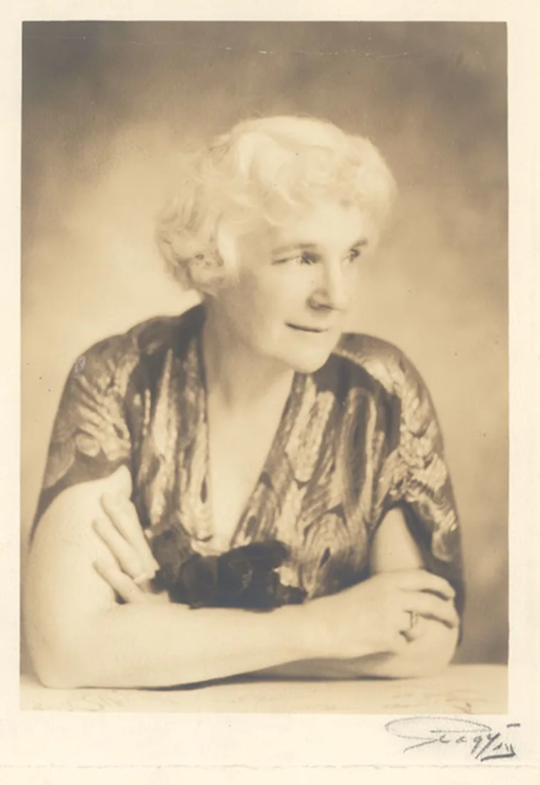 Person with curly hair sitting at a table, gazing sideways, wearing a patterned top. The image has a soft, sepia tone.