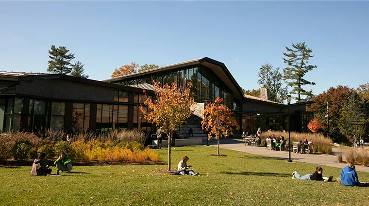 Modern building with large windows surrounded by trees. People sit and relax on the grass, enjoying a sunny day.