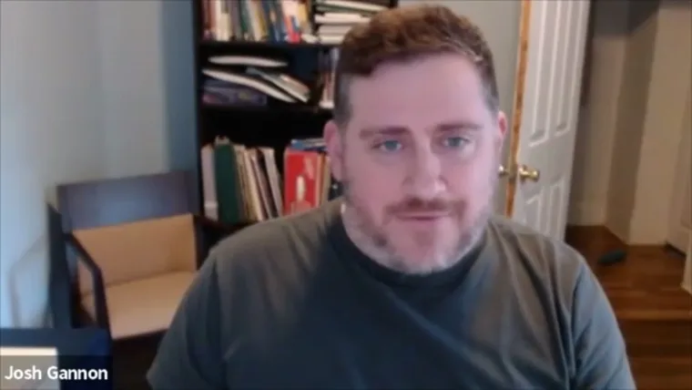 Person with short hair and beard in a room with a bookshelf and door in the background, wearing a gray shirt.