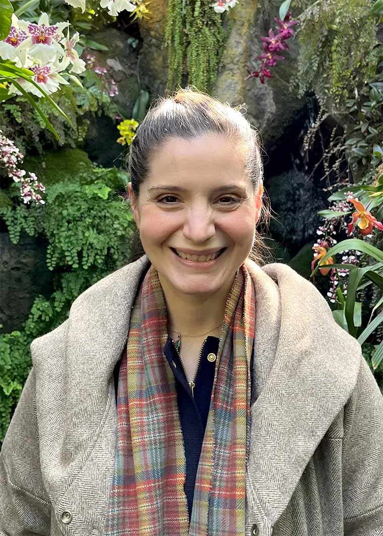 Person smiling in a garden filled with colorful flowers and greenery, wearing a patterned scarf and coat.