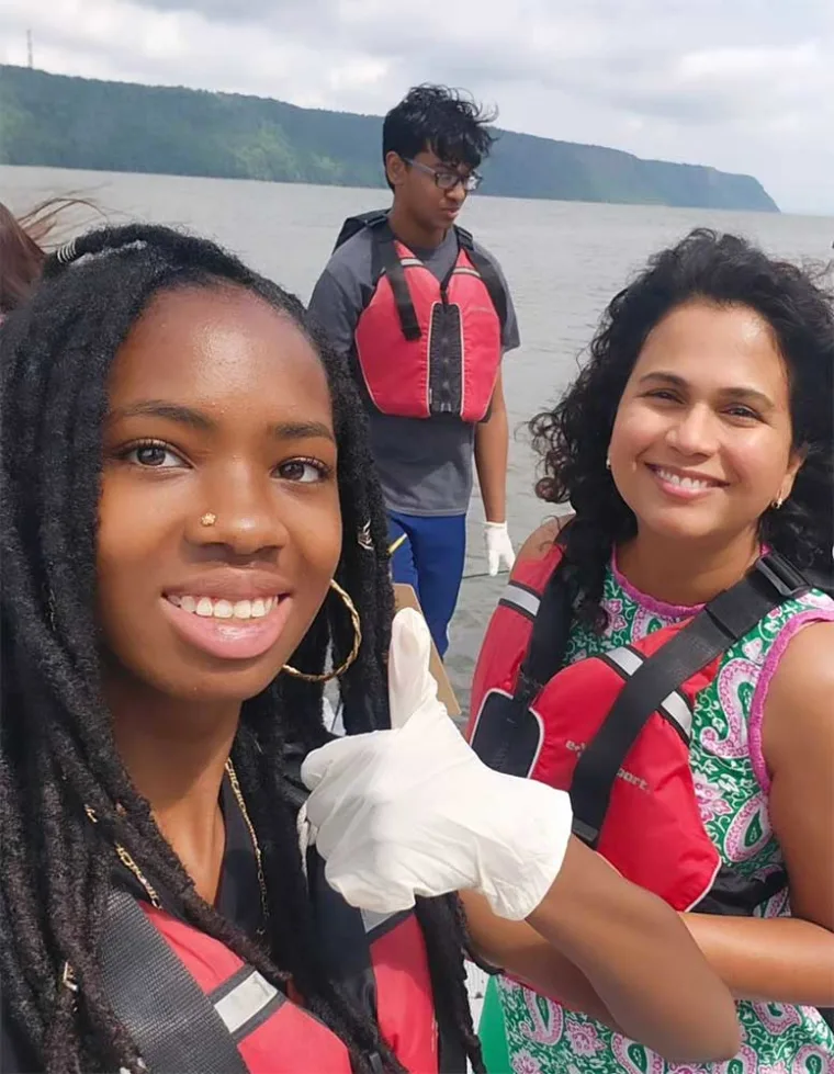 Three people in red life jackets stand on a boat with water and hills in the background, one giving a thumbs-up gesture.