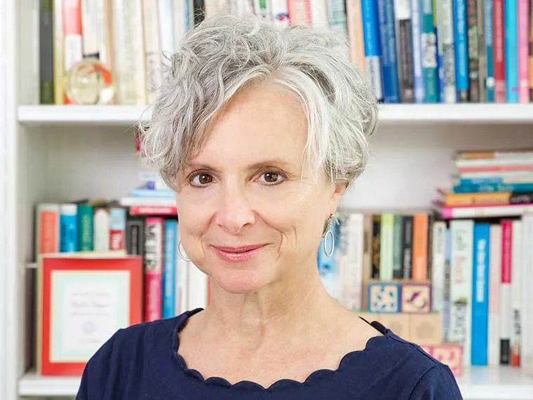 Person with short curly gray hair, wearing hoop earrings and a navy top, stands in front of a bookshelf filled with colorful books.