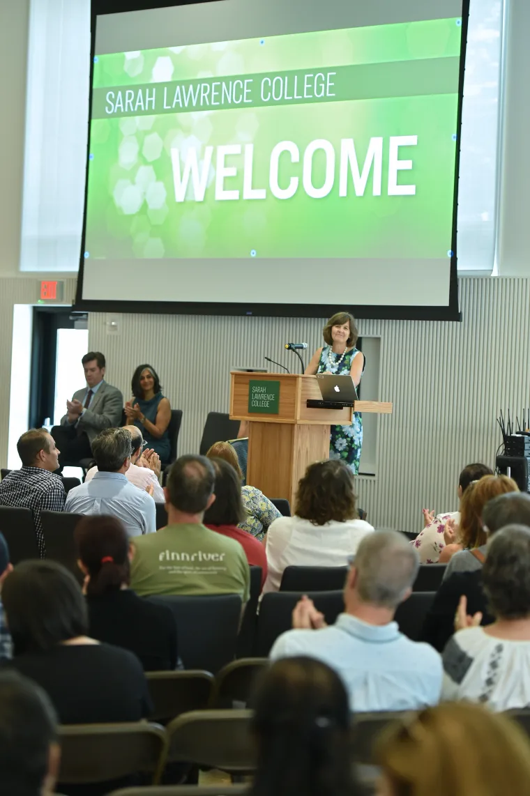 Person speaking at a podium on stage with a "Welcome" sign in the background. Audience seated and applauding.