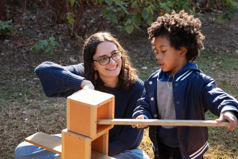 Person with glasses smiles at a child outdoors as they work together on a wooden project, surrounded by grass and plants.