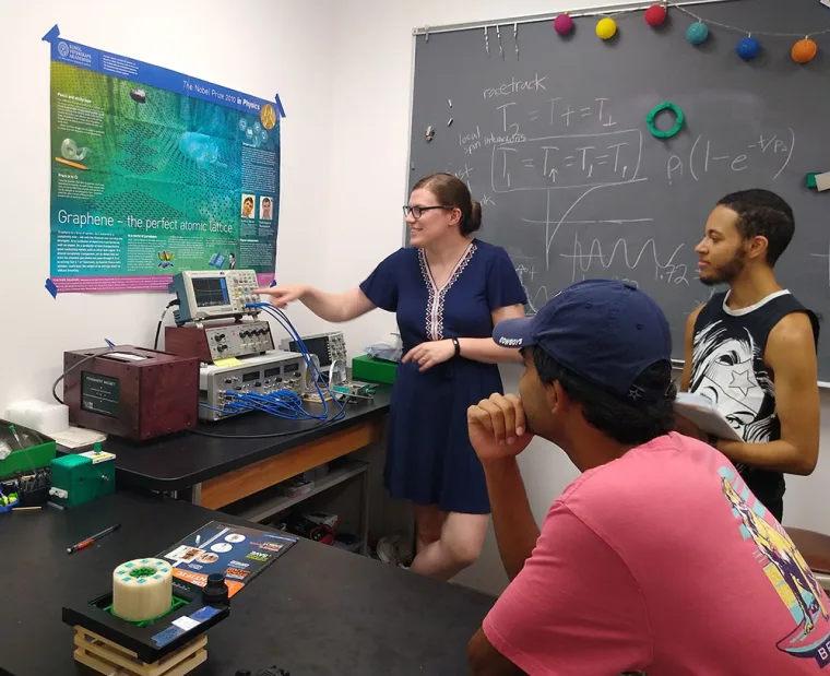 Three people in a lab, one pointing at equipment on a table, with a poster and chalkboard covered with equations in the background.