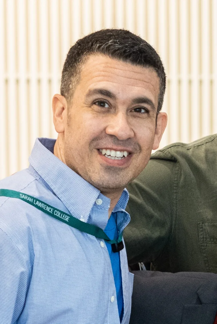 Person smiling, wearing a blue shirt and a lanyard with "Sarah Lawrence College" text, against a striped background.