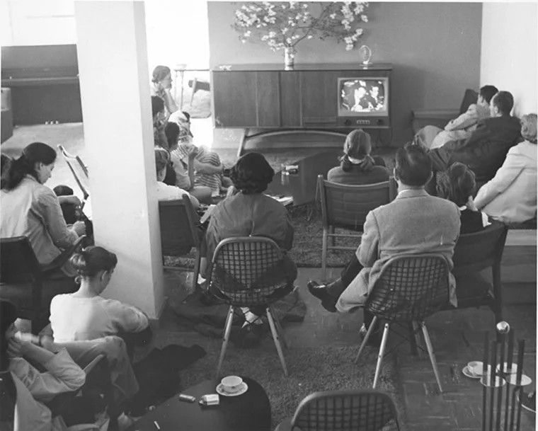 Group of people sitting and watching a vintage television in a living room. A vase of flowers is on top of the TV cabinet.