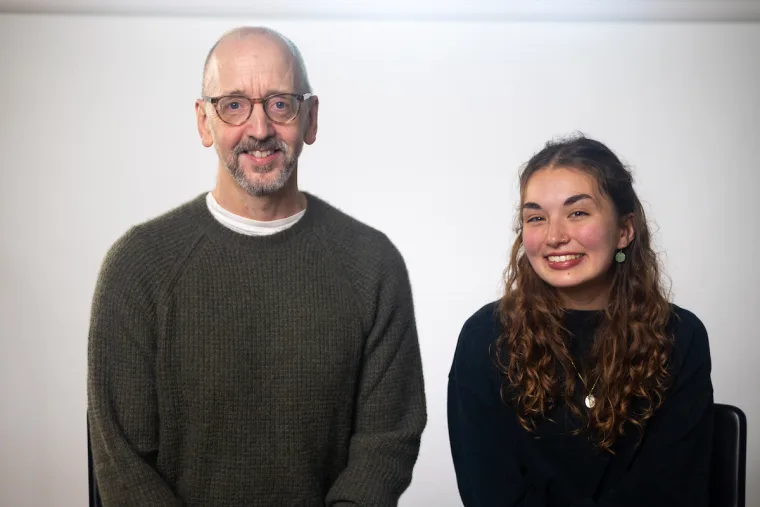 Two people sit side by side against a plain background, both smiling. One wears glasses and a sweater; the other has curly hair and a dark shirt.