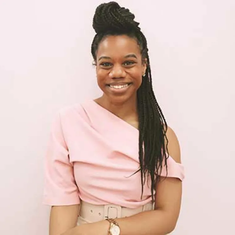 Person smiling, wearing a pink off-shoulder top, with long braided hair styled up, standing against a pale background.