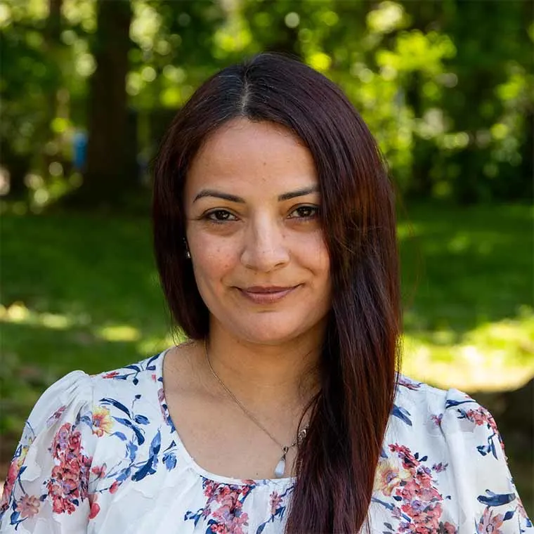 Person with long dark hair smiles slightly, wearing a floral top with a necklace. Lush green background suggests an outdoor setting.