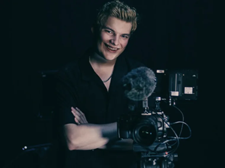 Person smiling, standing behind a professional camera with a microphone attached, against a dark background.