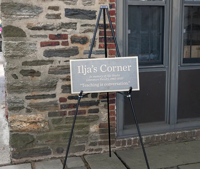 Sign on easel reads "Ilja’s Corner, In memory of Ilja Wachs, Literature Faculty, 1965–2022. 'Teaching is conversation.'" Set against a stone wall.