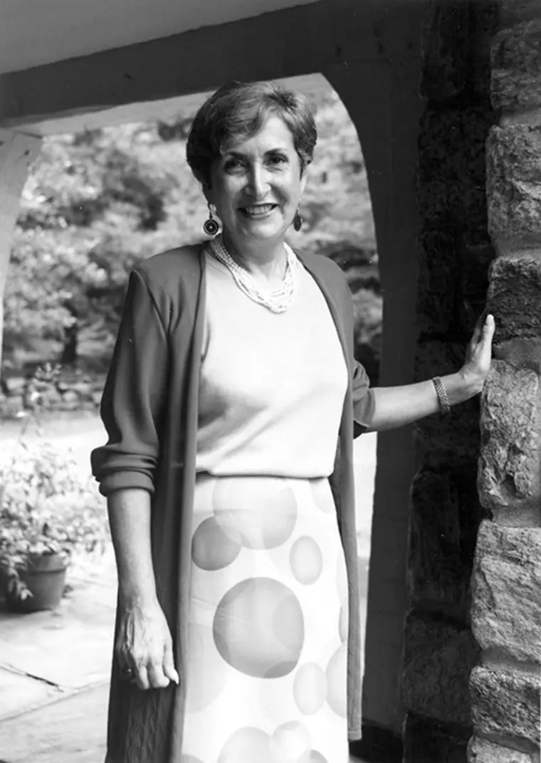 Person standing outdoors, smiling, wearing a cardigan and a polka dot skirt, with one hand resting on a stone wall. Trees in the background.