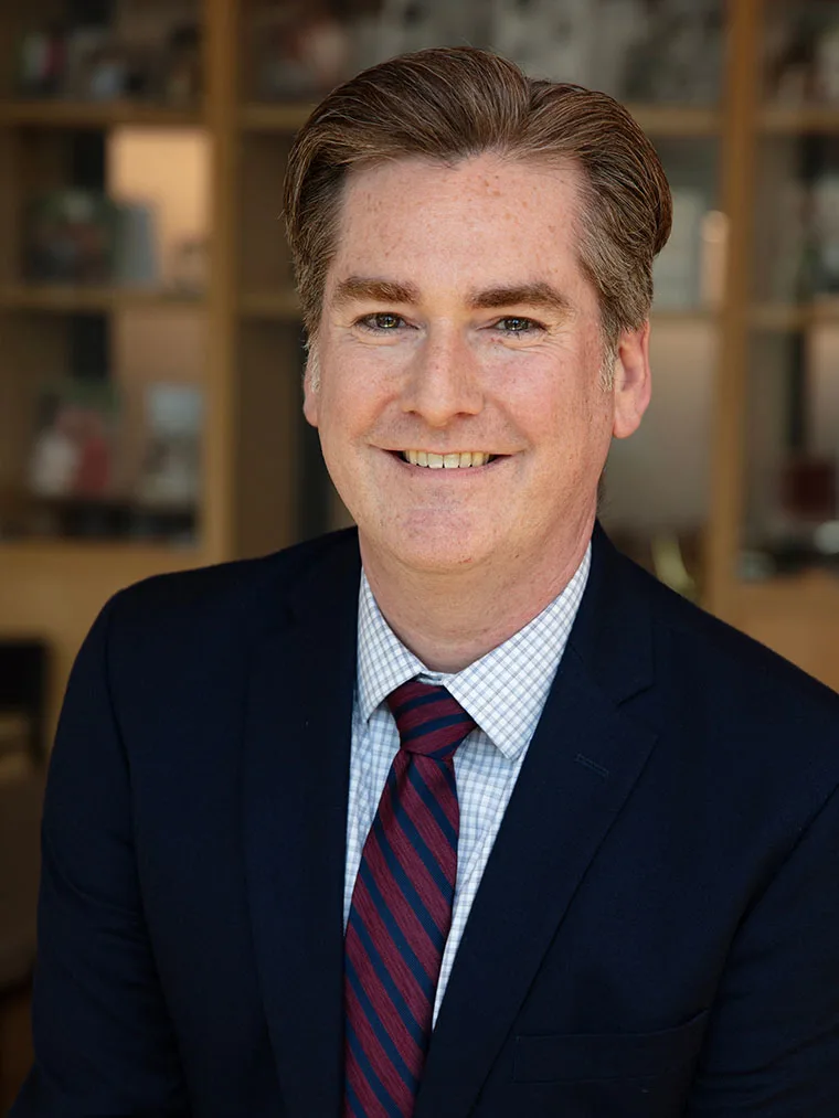 Person smiling, wearing a dark suit, checkered shirt, and red-striped tie, in an office setting with blurred shelves in the background.